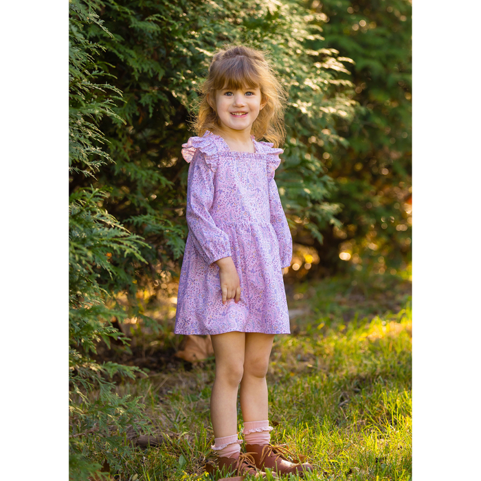 Young girl standing outdoors wearing a pink paisley print dress with long sleeves and ruffle trim, smiling in front of green foliage.