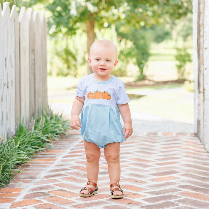 A toddler standing on a brick path outdoors, wearing a light blue romper dress with a pumpkin graphic on the chest and brown sandals.
