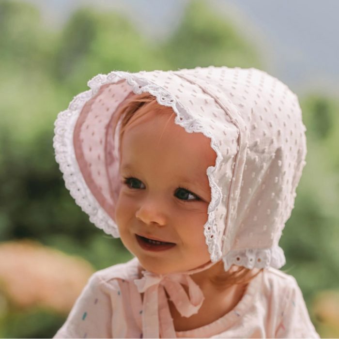 Toddler girl outdoors in a light pink clip dot bonnet with white lace trim and chin ties.