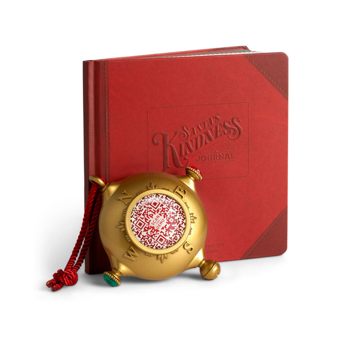 A golden-colored ornament with a red book titled 'Santa's Kindness Journal' and a red ribbon attached to it.
