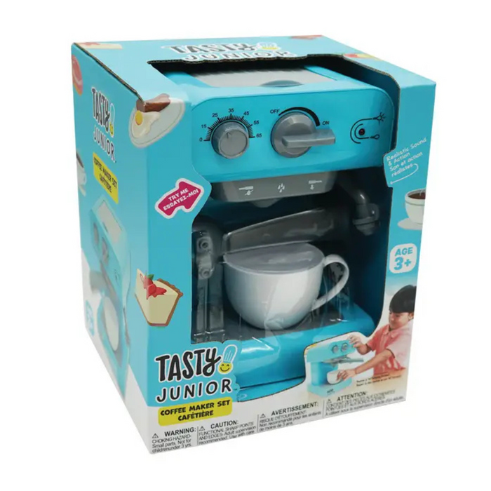 A young boy with blonde hair is sitting at a wooden table, playing with a light blue toy coffee maker set. He is turning a dial on the side of the machine, which has a white cup underneath the spout. The machine has "Tasty Junior" printed on the front.